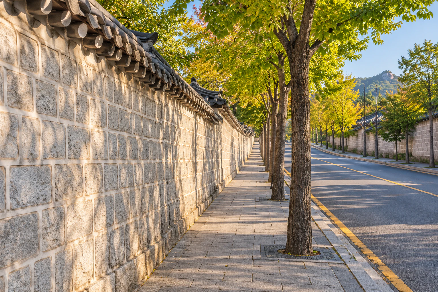把灰石上覆瓦的長牆放在左邊、往上走的窄人行道和行道樹。編輯圖片。
