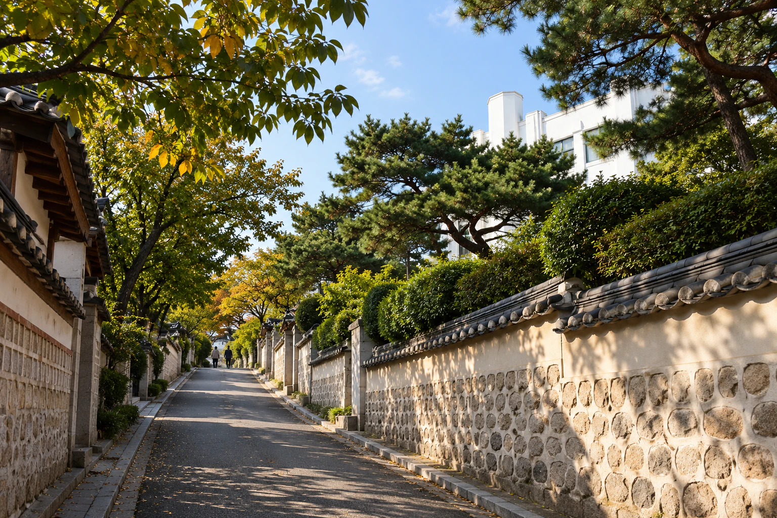 An uphill lane lined with walls and trees, part of a white building’s wall showing over the fence. Editorial image.