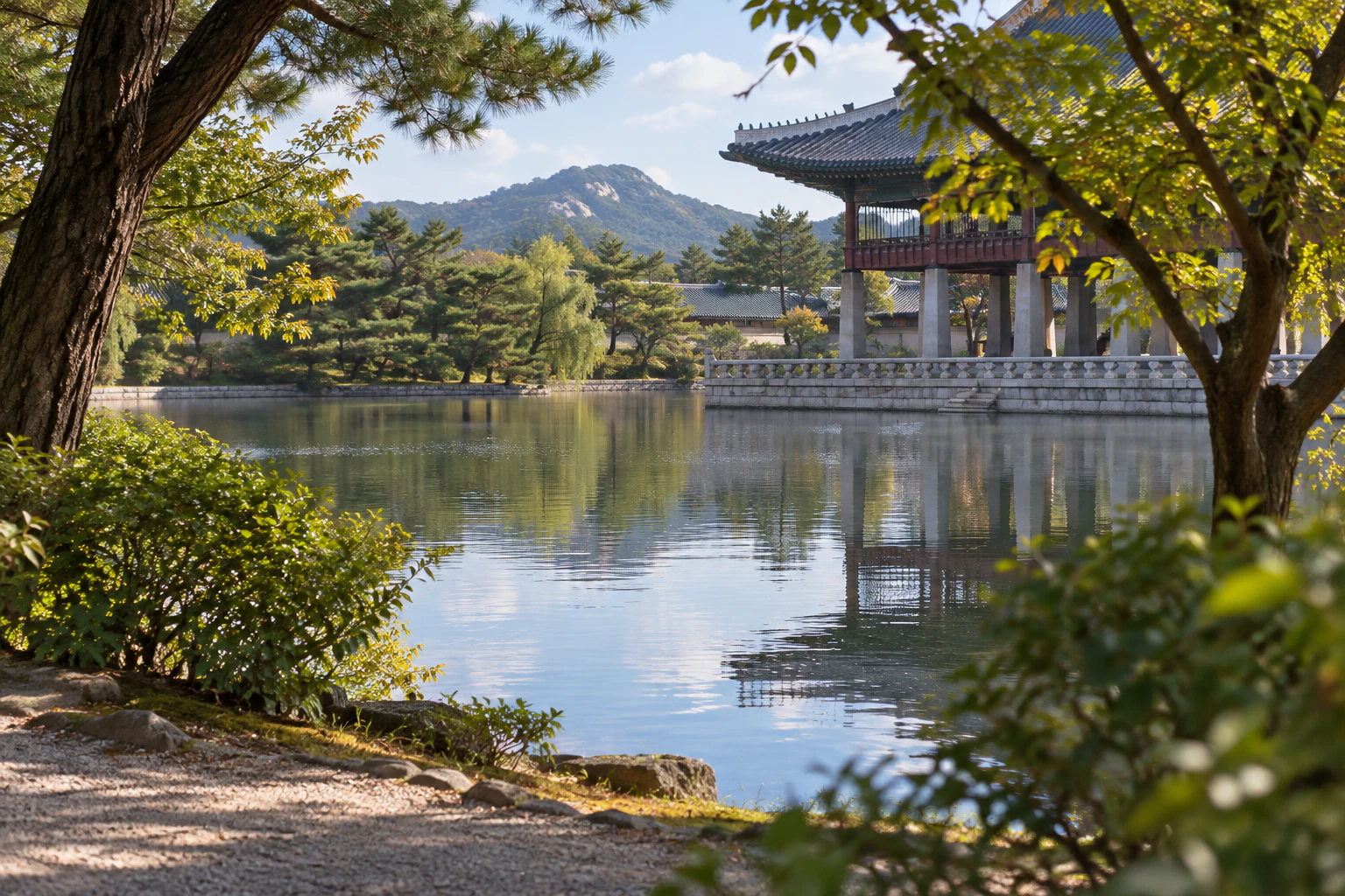 A pond glimpsed first between trees along a path, part of a pavilion behind it. Editorial image.