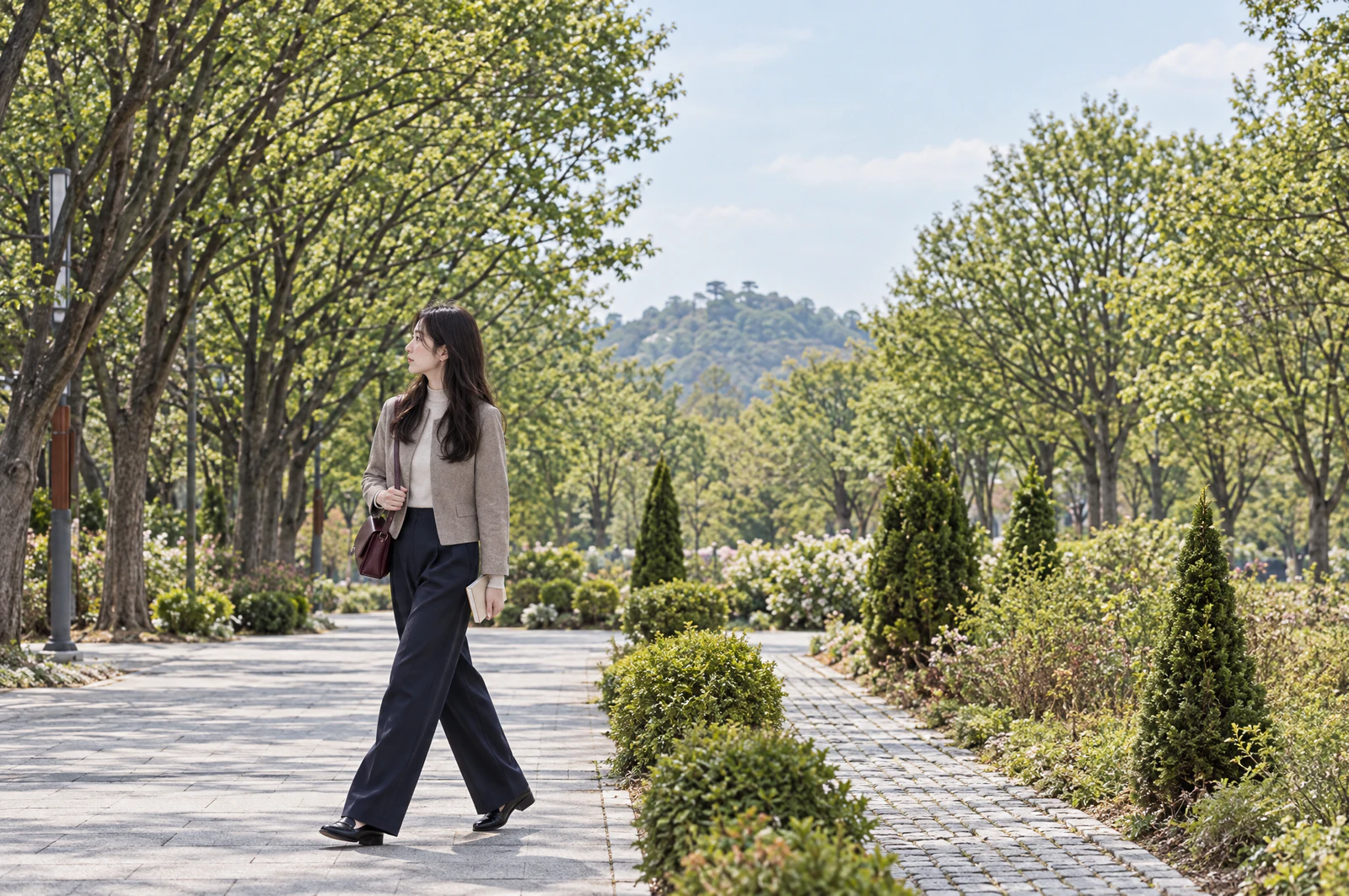A woman in a short taupe jacket with a burgundy crossbody bag walks along a tree-lined park path, looking towards the woods at her side. Editorial image.