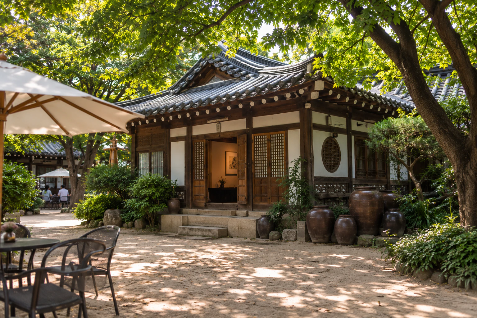 The front of a tiled-roof hanok across the sandy courtyard, timber posts, white walls and a round lattice window, onggi jars in front, a white parasol and iron table in the foreground. Editorial image.