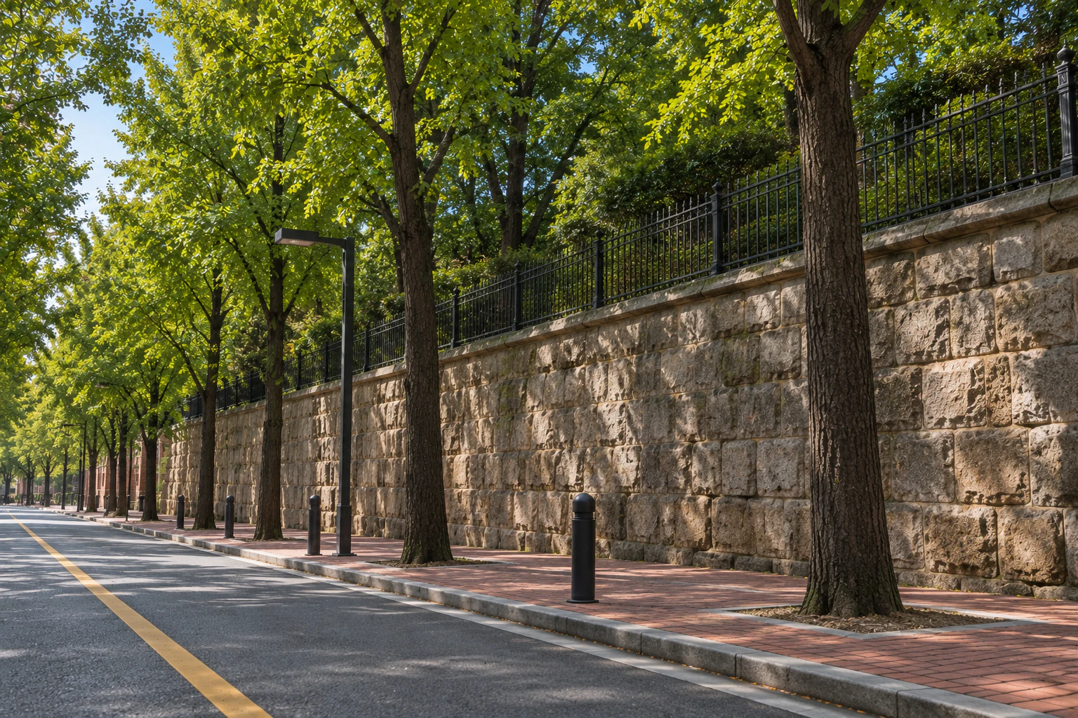 A high retaining wall of stacked stone with an iron railing on top, trees behind it and ginkgo street trees in front.