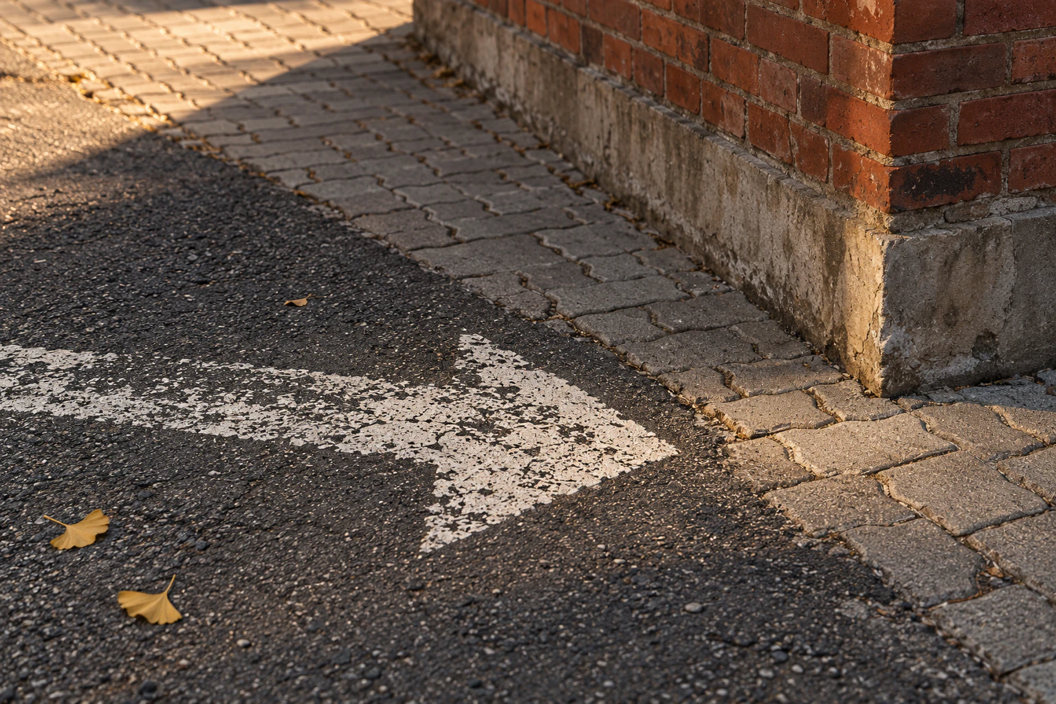 A worn white painted arrow on dark asphalt pointing toward the corner of a red-brick wall in a Seongsu back lane, a few ginkgo leaves on the pavers, no people