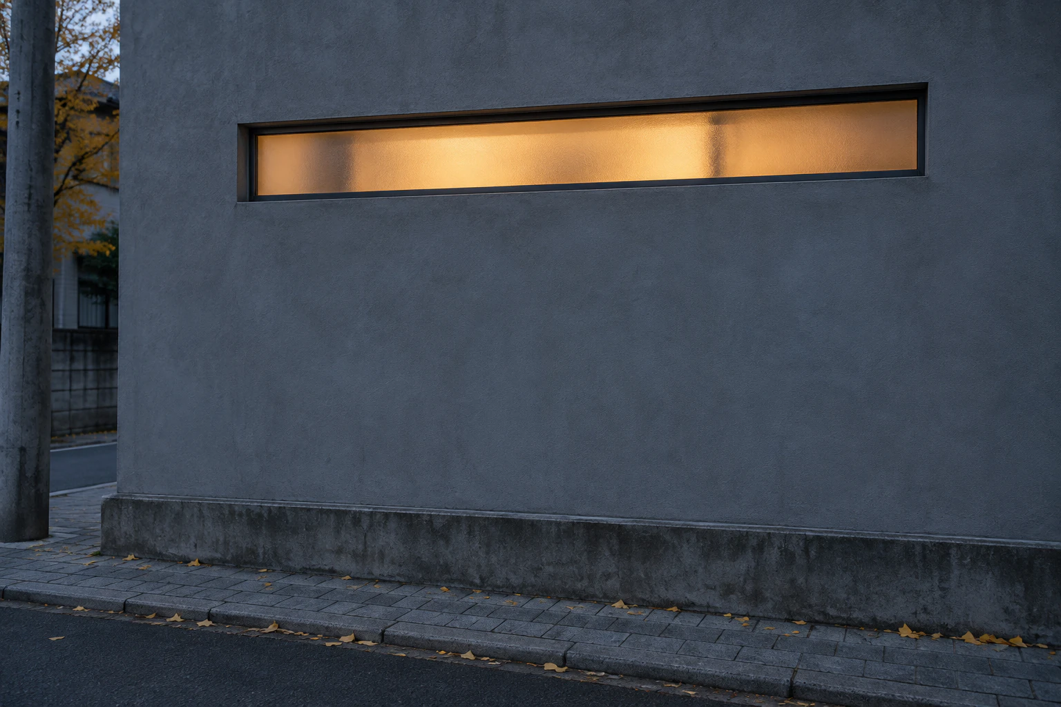 A plain grey rendered wall of a small building in a Seongsu back lane at dusk with one narrow frosted window glowing warm near the top, no door, no sign, a grey utility pole at the left edge