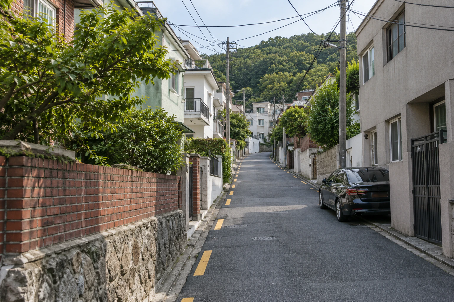An asphalt residential street runs uphill between a red-brick wall on a stone base and low-rise homes. Overhead cables cross the view, a car is parked on the right, and a wooded slope rises behind the buildings. No people.