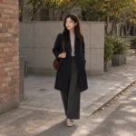 Rina walks along a brick alley in a navy coat, grey turtleneck, charcoal trousers and an oxblood shoulder bag.