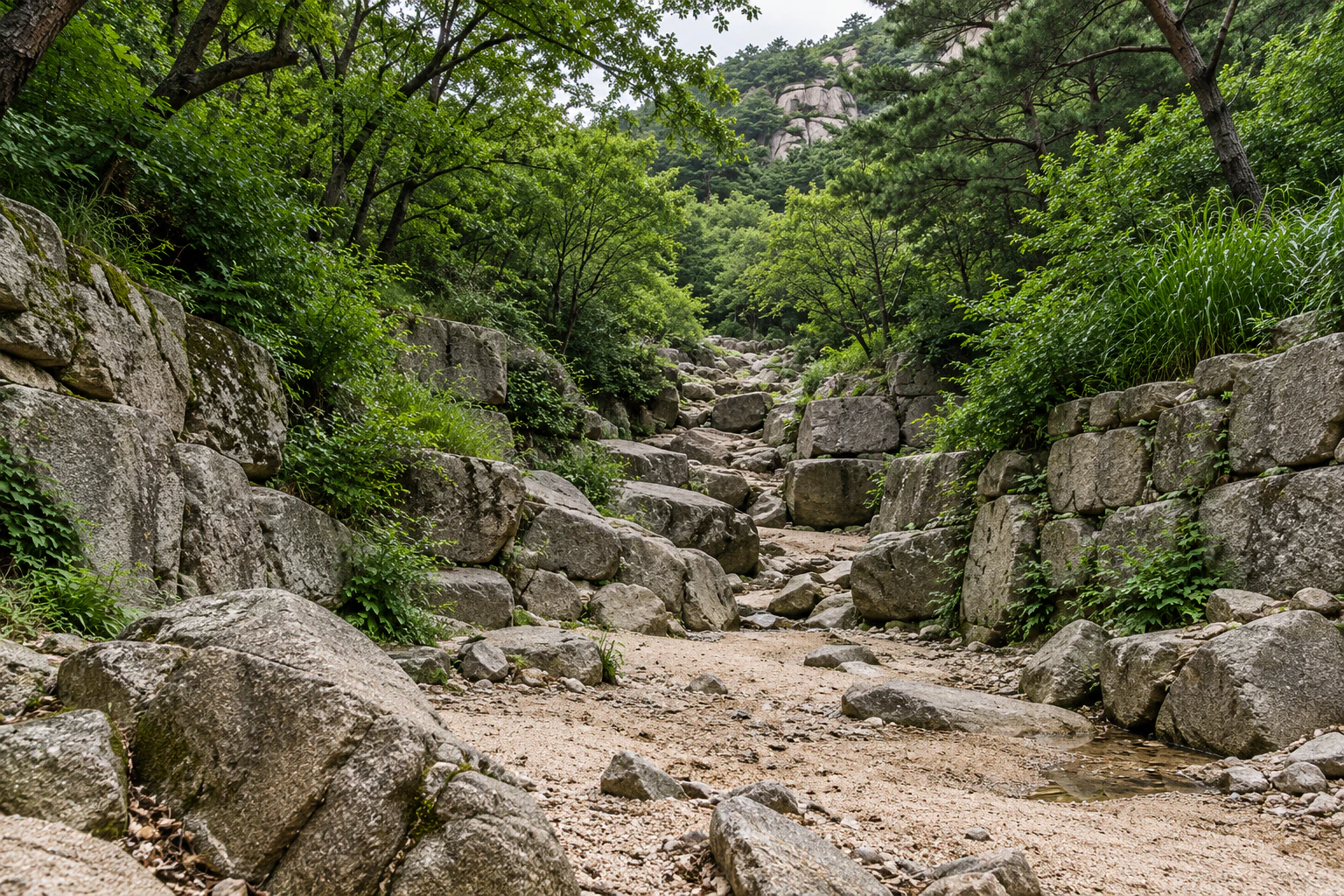 A nearly dry streambed of sand and stones between stone retaining walls and dense trees. No people. Editorial image.