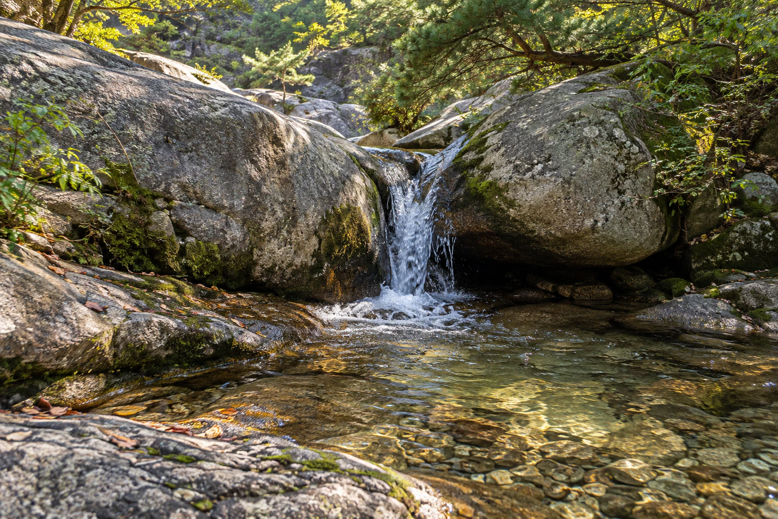 A small cascade falling between granite boulders into a clear pool with stones visible on the bottom. No people. Editorial image.