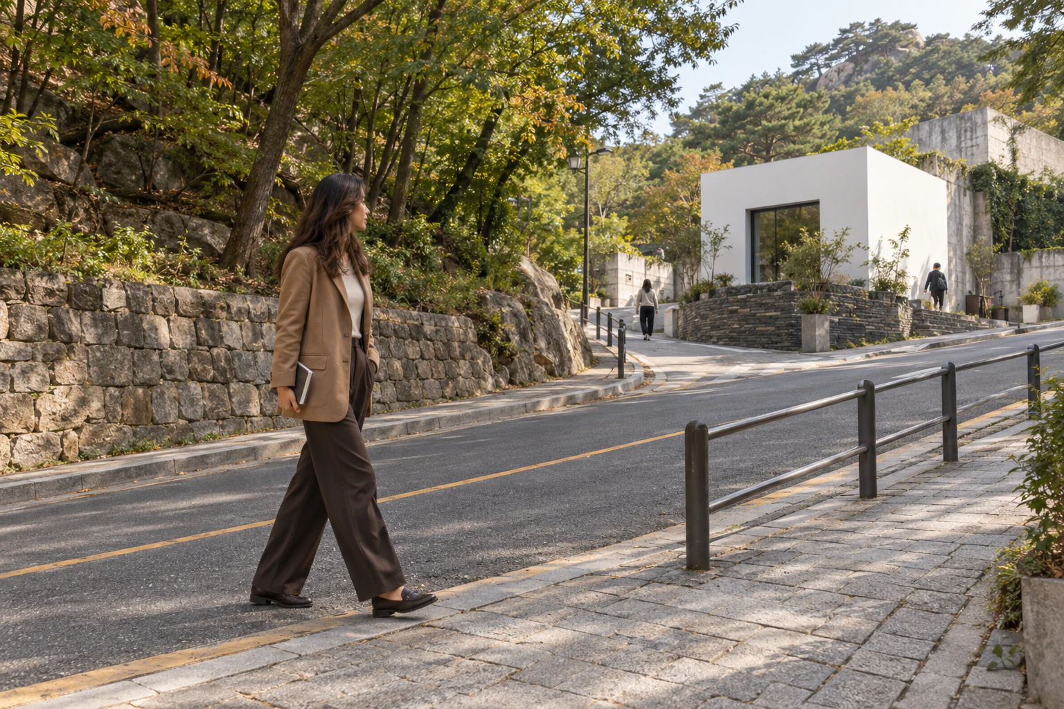 A woman with long, wavy brown hair in a camel blazer walks up a sloping pavement in profile, looking across the road. Editorial image.