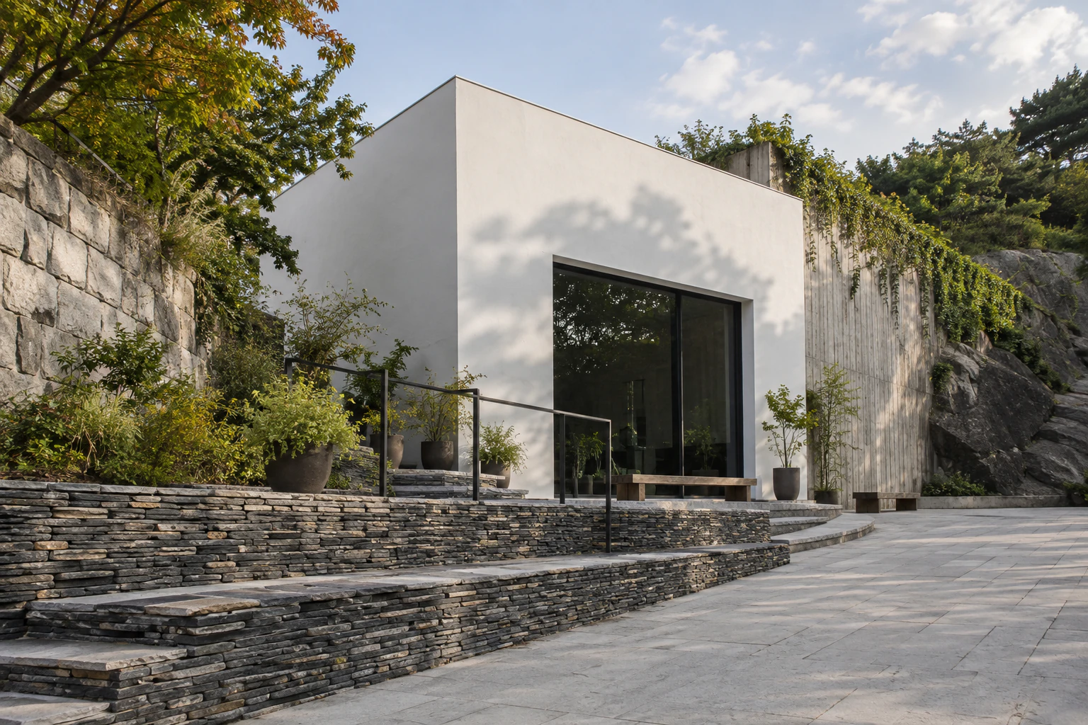 A crisp white box-shaped building beside rough bedrock and a stone retaining wall, with low terraces of thinly stacked stone in front. No people. No lettering. Editorial image.