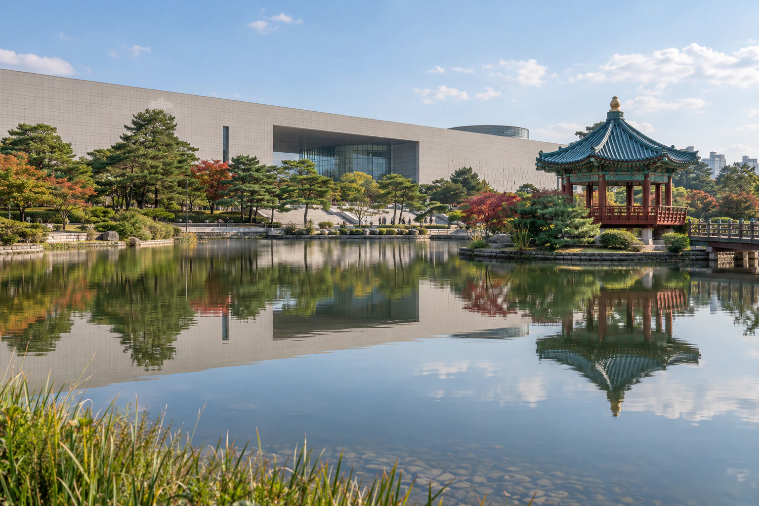 A wide pond reflecting a pale stone building and a pavilion with a green roof. People small and distant. No lettering. Editorial image.