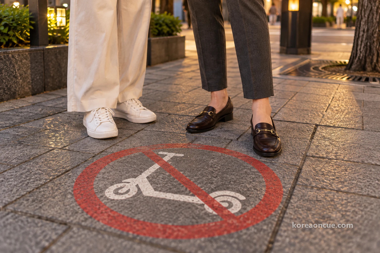 A low-angle close-up of a kick-scooter pictogram inside a red circle with a slash on the evening pavement, beside two pairs of shoes, including white sneakers. Editorial image.