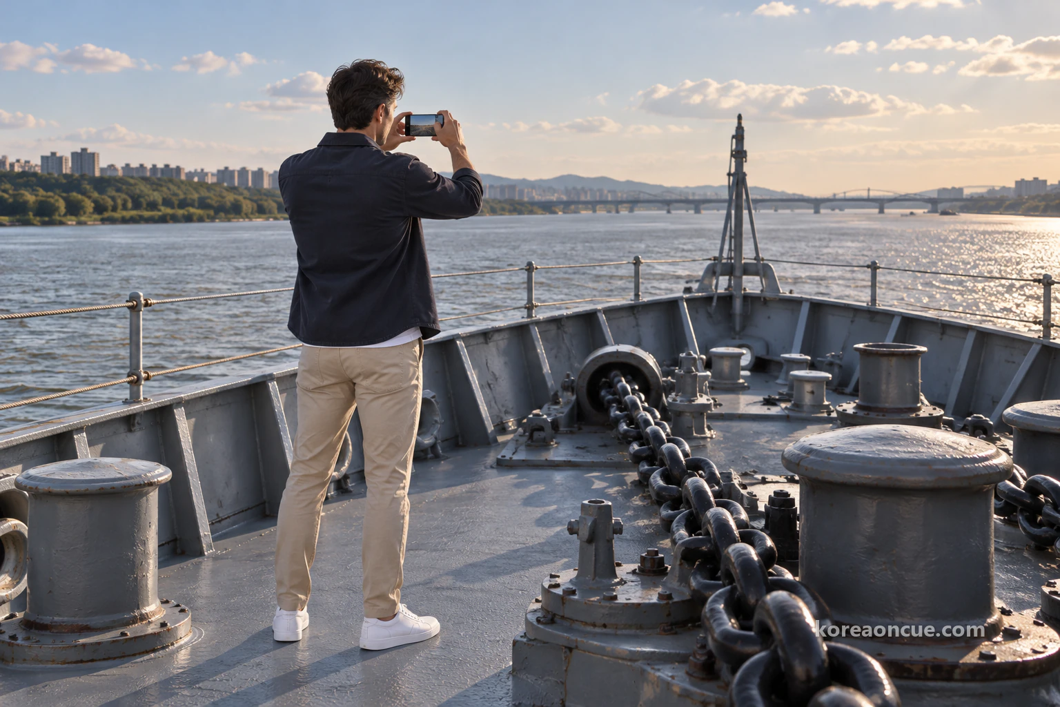 Ethan seen from behind photographing the river with his phone on a frigate’s bow deck beside a thick anchor chain and deck fittings in afternoon light. Editorial image.