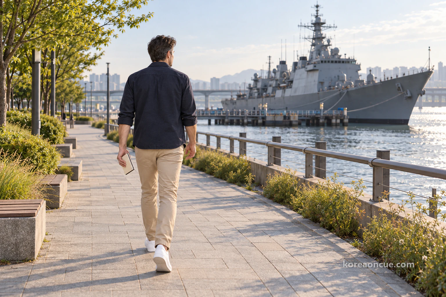 Ethan seen from behind walking along a riverside park path with a notepad in one hand, a long grey naval ship moored at the bank in the distance. Editorial image.