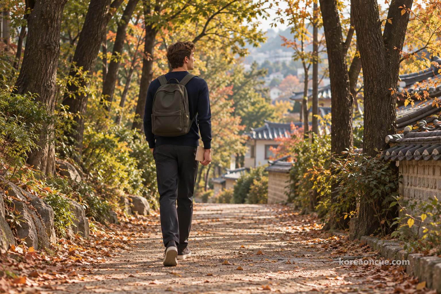 Noah seen from behind walking alone along a forest trail lined with tall trees on an afternoon, notebook in hand, low rooftops showing between the trunks and fallen leaves on the path. Editorial image.