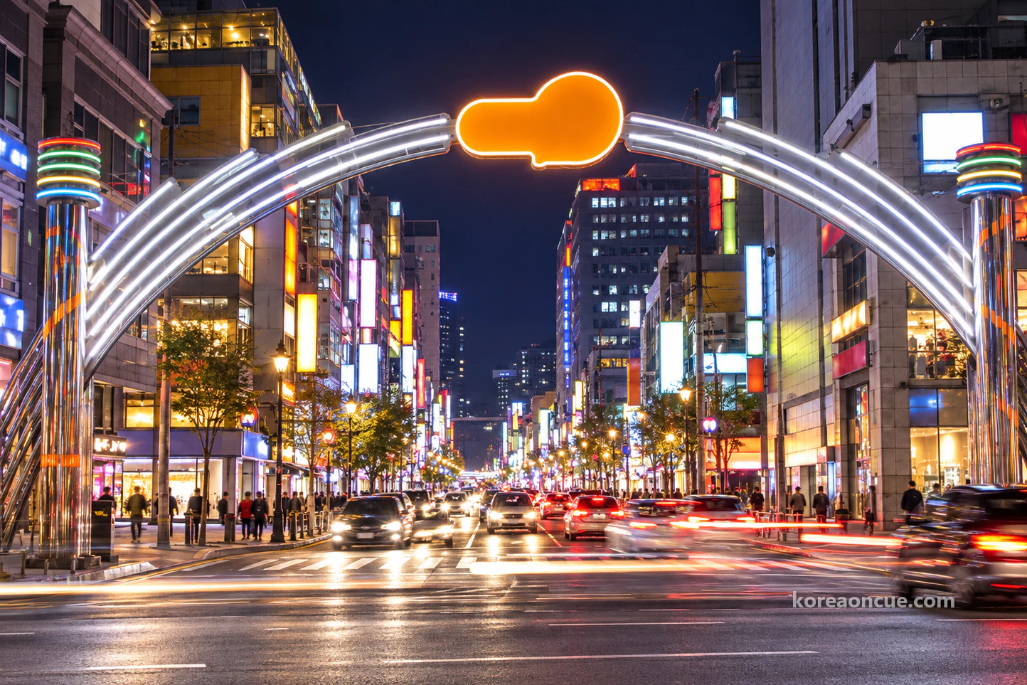 A large arch of two white light bands spanning a road beneath a deep navy sky, rising from striped pillars to a glowing orange speech-bubble shape, commercial buildings lit on every floor, long car light trails and a few small pedestrians at the kerb. Editorial image.