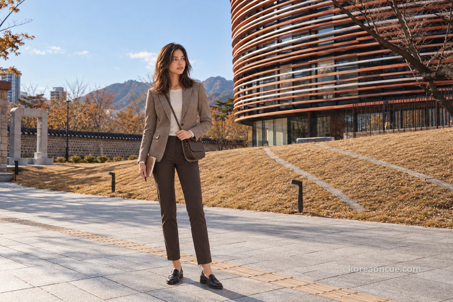 Full-length view of Elena holding a notebook and turning towards a building on the left in a courtyard between low museum buildings on an autumn afternoon, with a rounded orange-and-white banded wall and a low boundary wall. Editorial image.