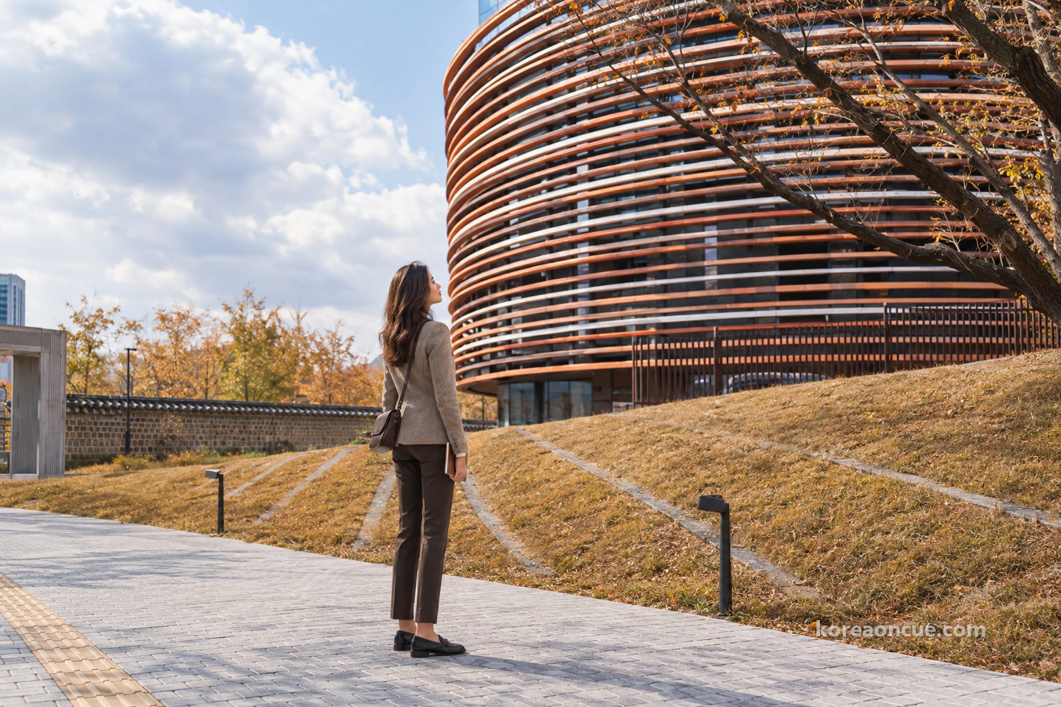 Elena seen from behind at an angle, her profile visible as she holds a folded notebook and looks up from below a brown grass mound edged with stone at a rounded museum facade with orange and white horizontal bands, a stone gate frame and low wall to the left in clear autumn afternoon light. Editorial image.