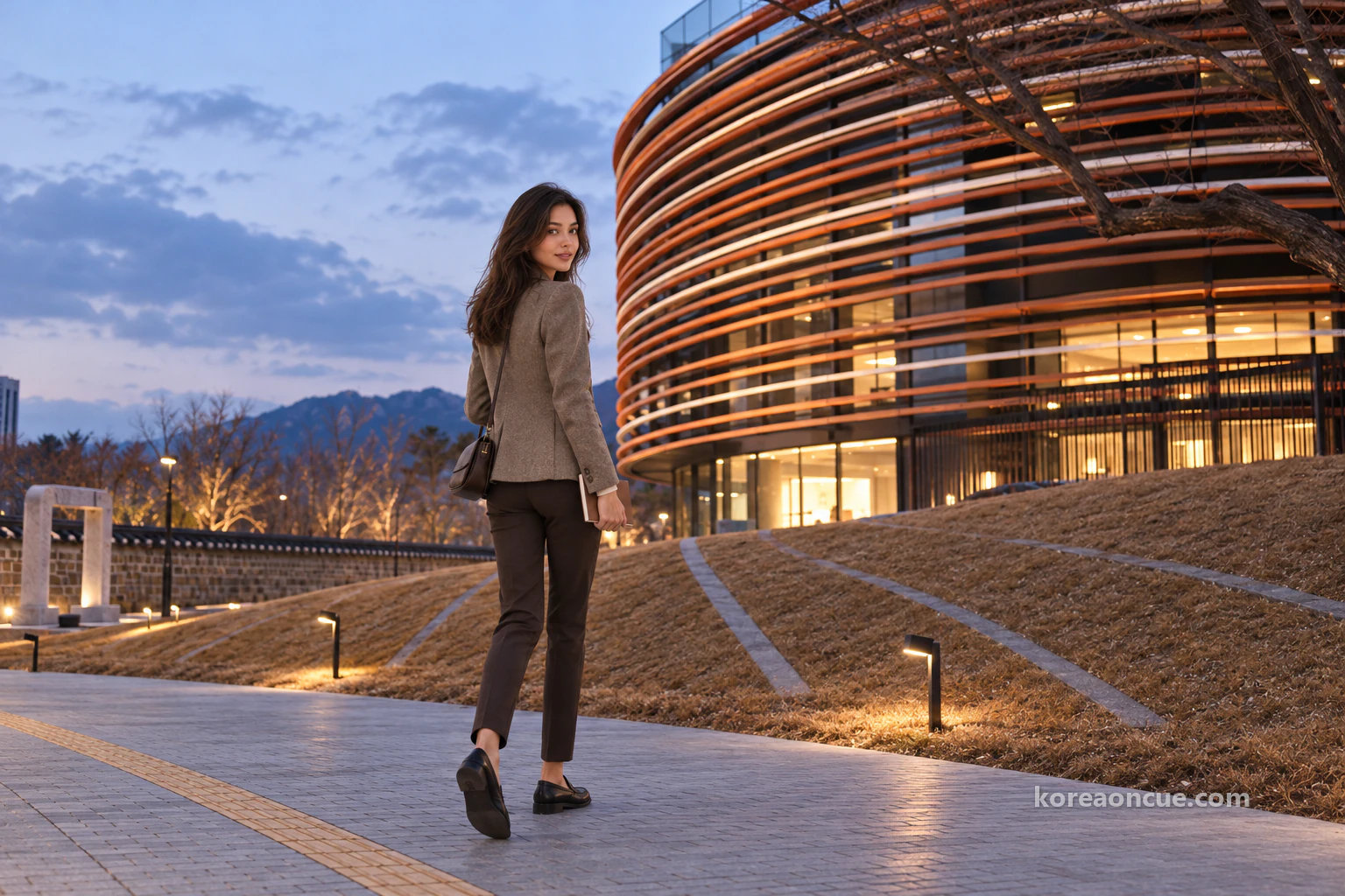 Rear three-quarter view of Elena carrying a small notebook across a museum courtyard at blue hour towards a lit building, her face turned slightly with a smile, a rounded orange-and-white banded facade with warm windows, a lawn mound edged with stone, a low wall and stone gate frame to the left, and a distant mountain ridge. Editorial image.