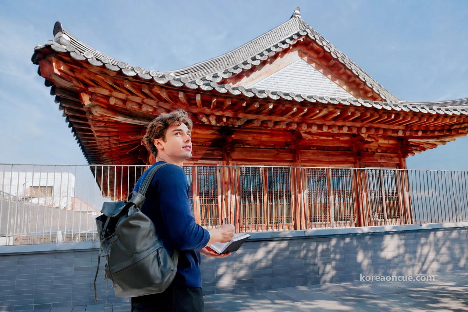 A large modern hanok-style building with a grey hipped-and-gabled roof, red-brown wooden brackets and wooden-framed upper windows under a deep blue late-autumn sky, Noah at the left holding an open notebook and pen and looking up from the pavement, seen full-length from behind at an angle, a low tiled roof and thin metal railing in front. Editorial image.