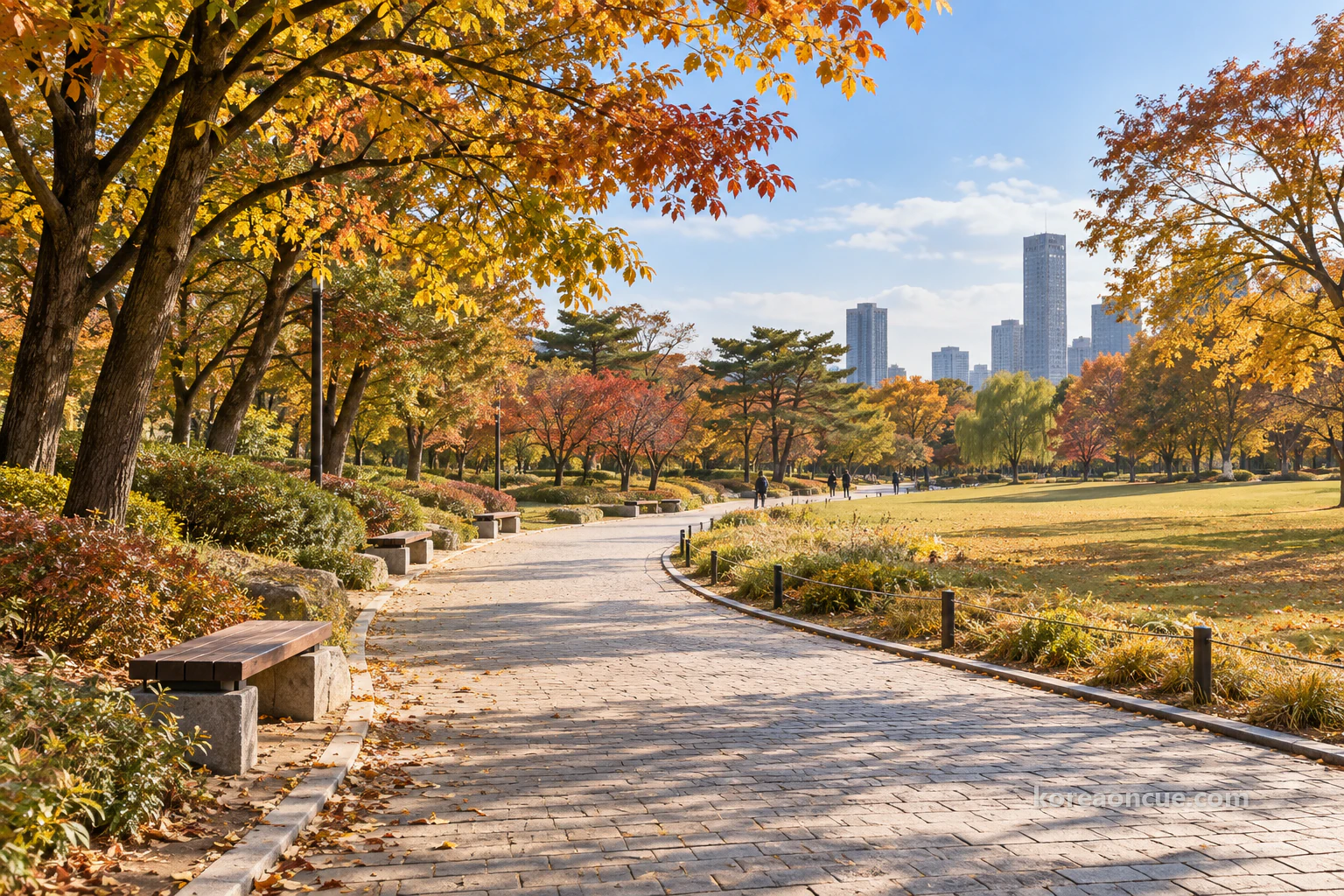 A broad park path curving gently between wide lawns in autumn sunlight, large trees beginning to change colour, low benches along the edge and a few tiny walkers in the distance. Editorial image.