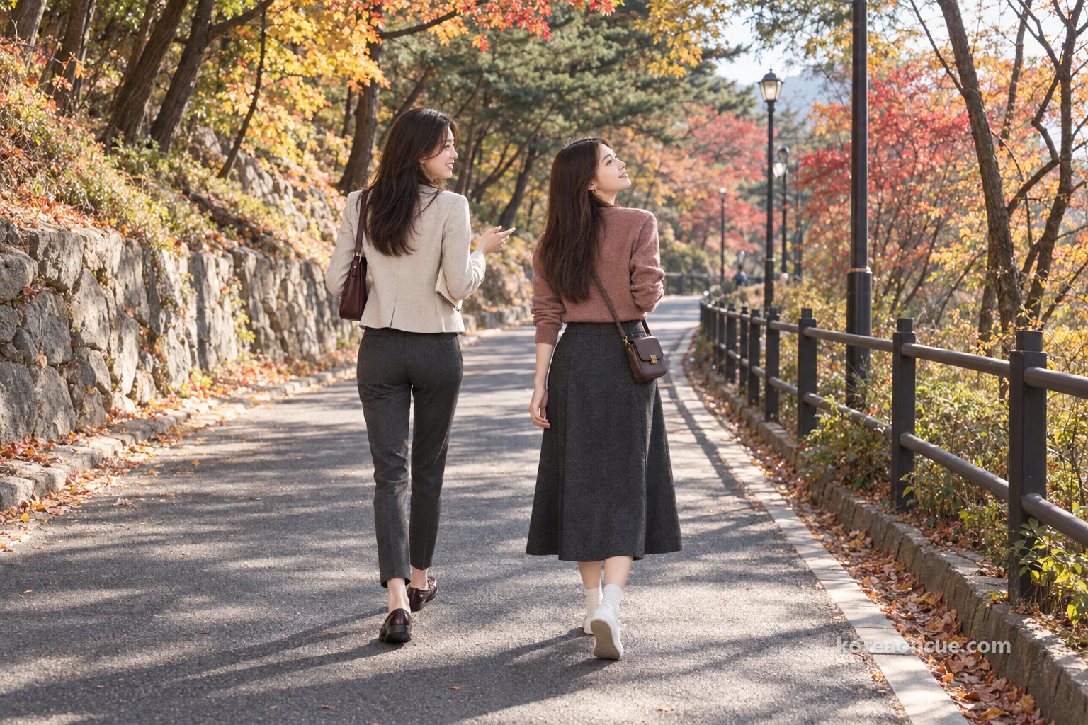 Rina and Mali seen from behind walking side by side along a wide, car-free walking road in slanting morning light, Rina with her notebook under her arm turning to talk to Mali, Mali looking up at the branches, autumn trees and pines on the slopes, a low railing and a row of street lamps stretching ahead. Editorial image.