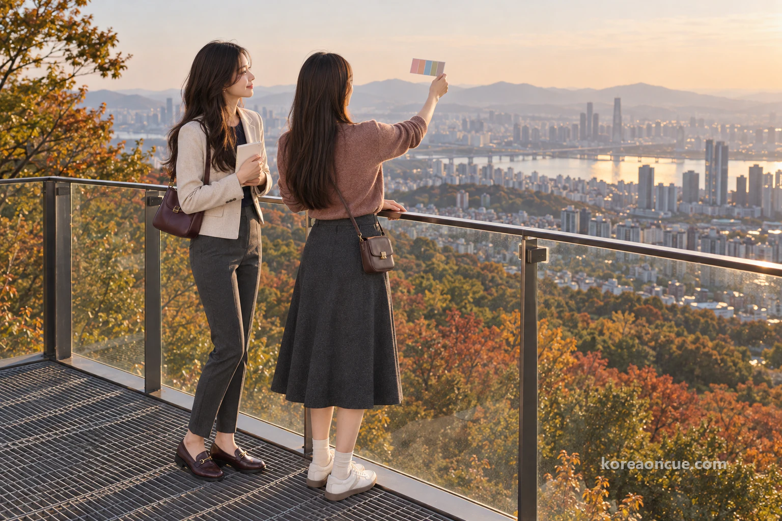 Rina and Mali seen from behind at the end of a viewing deck with a steel grating floor and a clear glass railing in low late-afternoon sun, Mali holding a colour card up to the sky over the city, Rina hugging her notebook and following her fingertip, autumn forest below and a hazy Seoul and mountain ridges in the distance. Editorial image.