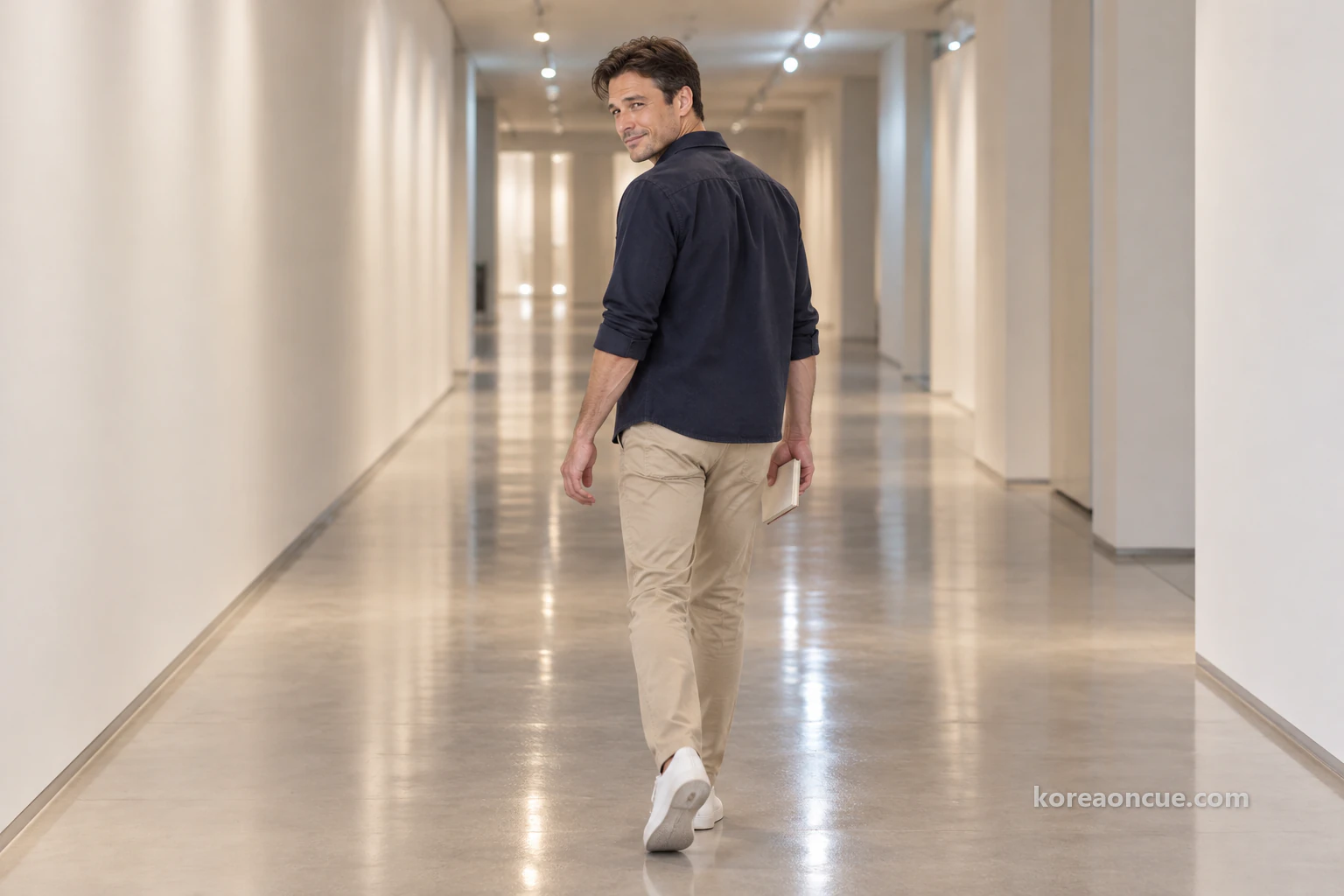 Ethan seen from behind walking slowly down a long, quiet white corridor, turning his head slightly to look back, ceiling lights reflected in long streaks on the floor. Editorial image.