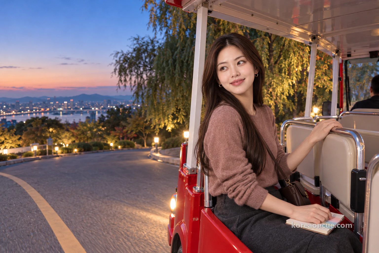 Just after sunset under a deep blue sky, Mali on a rear seat of a small red open-sided electric shuttle going down an asphalt slope beside willows with its headlights on, turning to look back at the sky above the hill with a quiet, satisfied smile, the edges of colour cards showing from the memo pad on her lap. Editorial image.