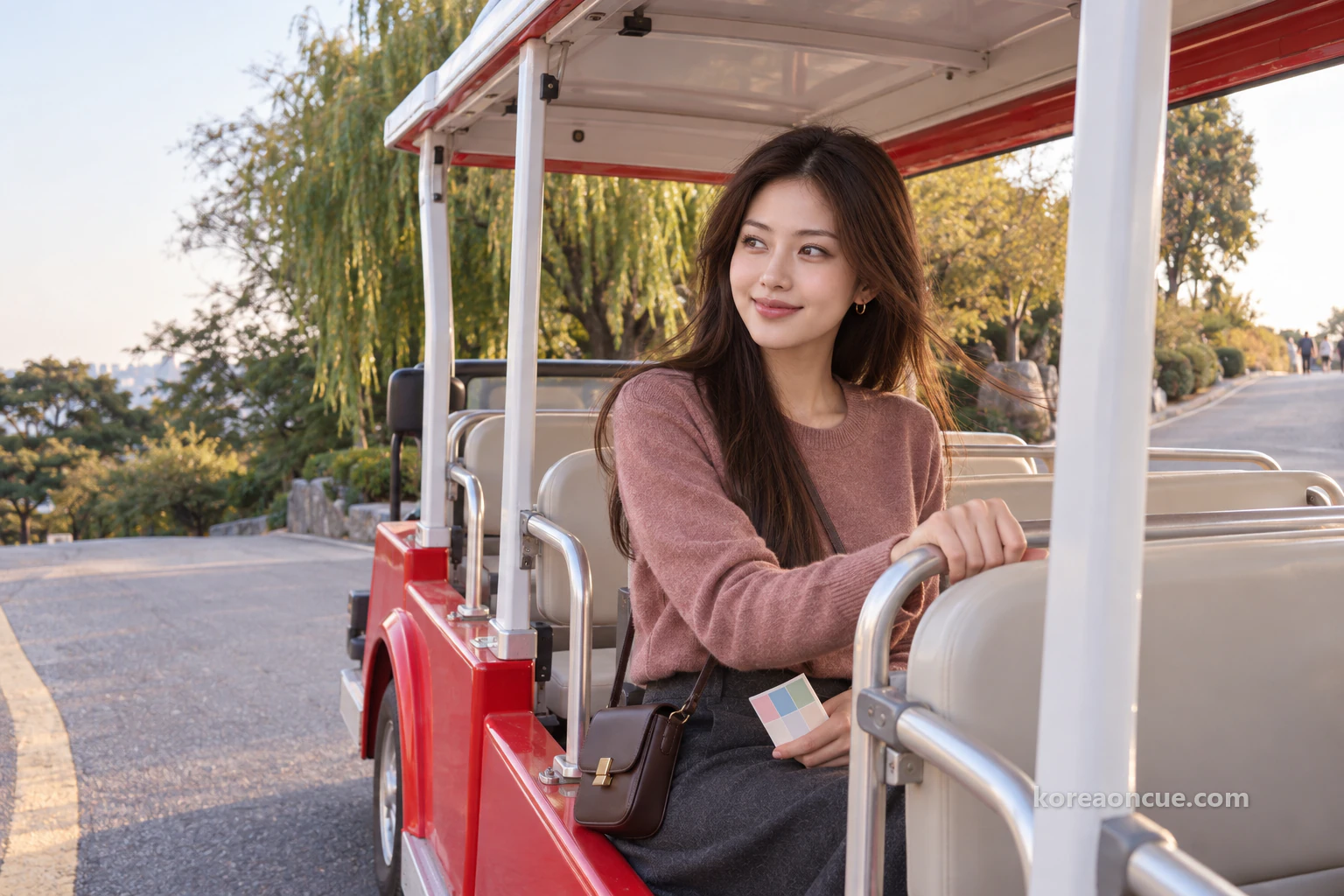 Mali in profile on a rear seat of a small red open-sided electric shuttle climbing an asphalt slope past drooping willows in late afternoon, looking out with an excited look, one hand on a silver handrail and a single colour card on her lap, the breeze lifting her hair. Editorial image.