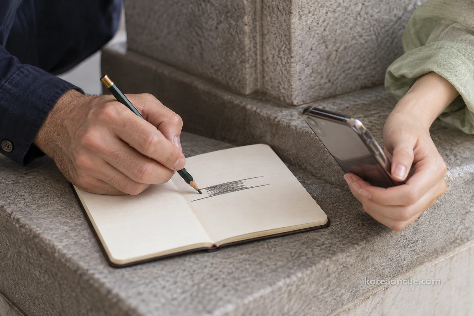 Close-up of a hand drawing two horizontal pencil lines across a small notebook on a grey ledge and shading where they overlap, another hand at the edge of the frame holding a phone towards the page. Editorial image.