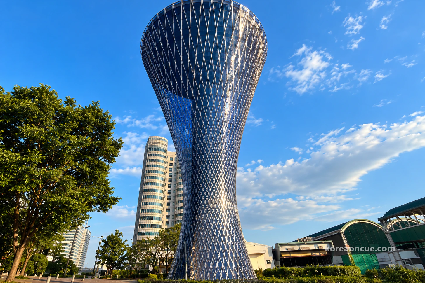 A tall hourglass-shaped tower wrapped in a shining silver lattice mesh under a clear blue sky, green trees and a low building with a curved green roof in front and a tall apartment block behind. Editorial image.