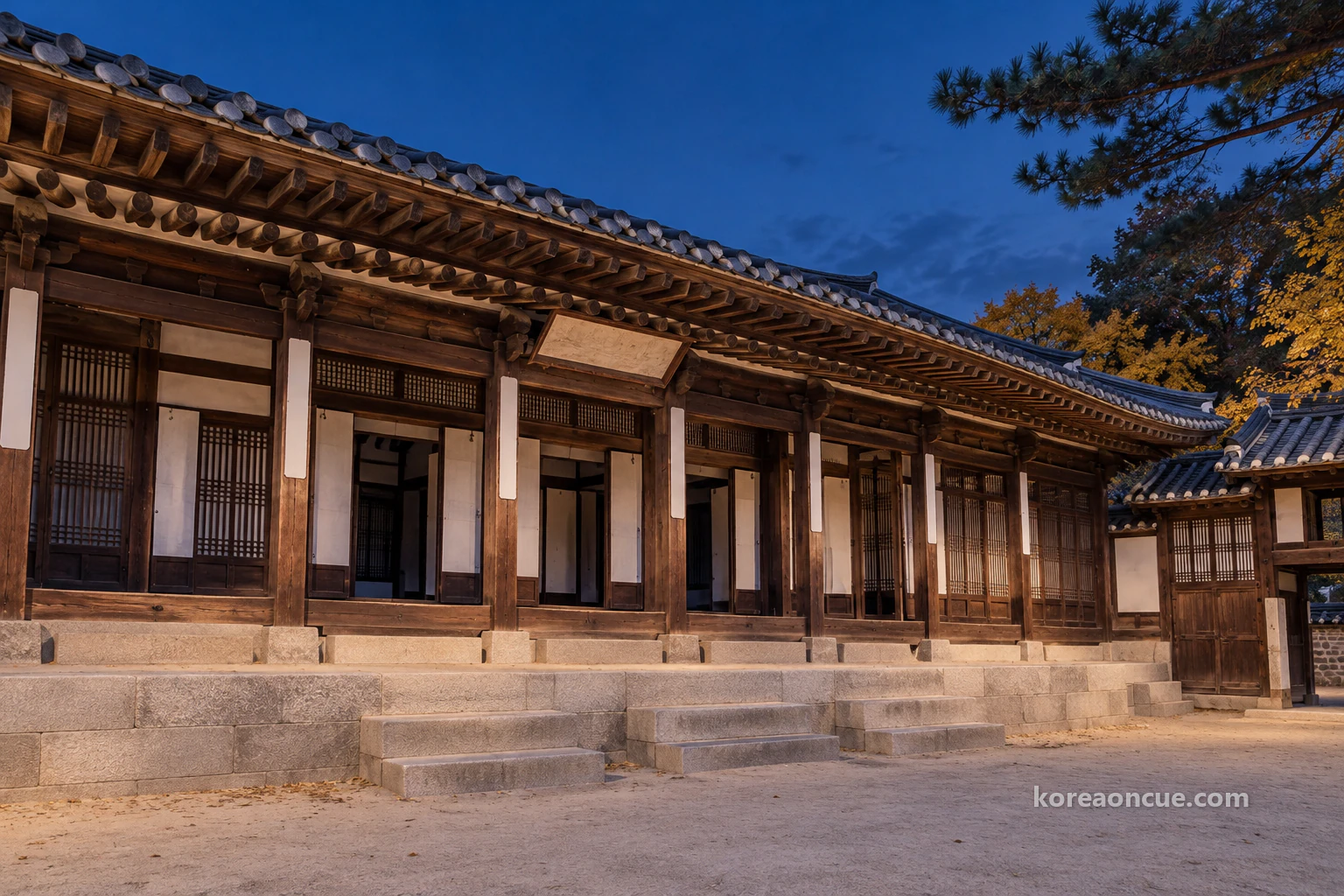 A long single-storey hanok front under a deep blue sky just after sunset, a broad stone platform with a central flight of steps, the double-layered tiled eaves in silhouette against the sky and a quiet, empty yard. Editorial image.