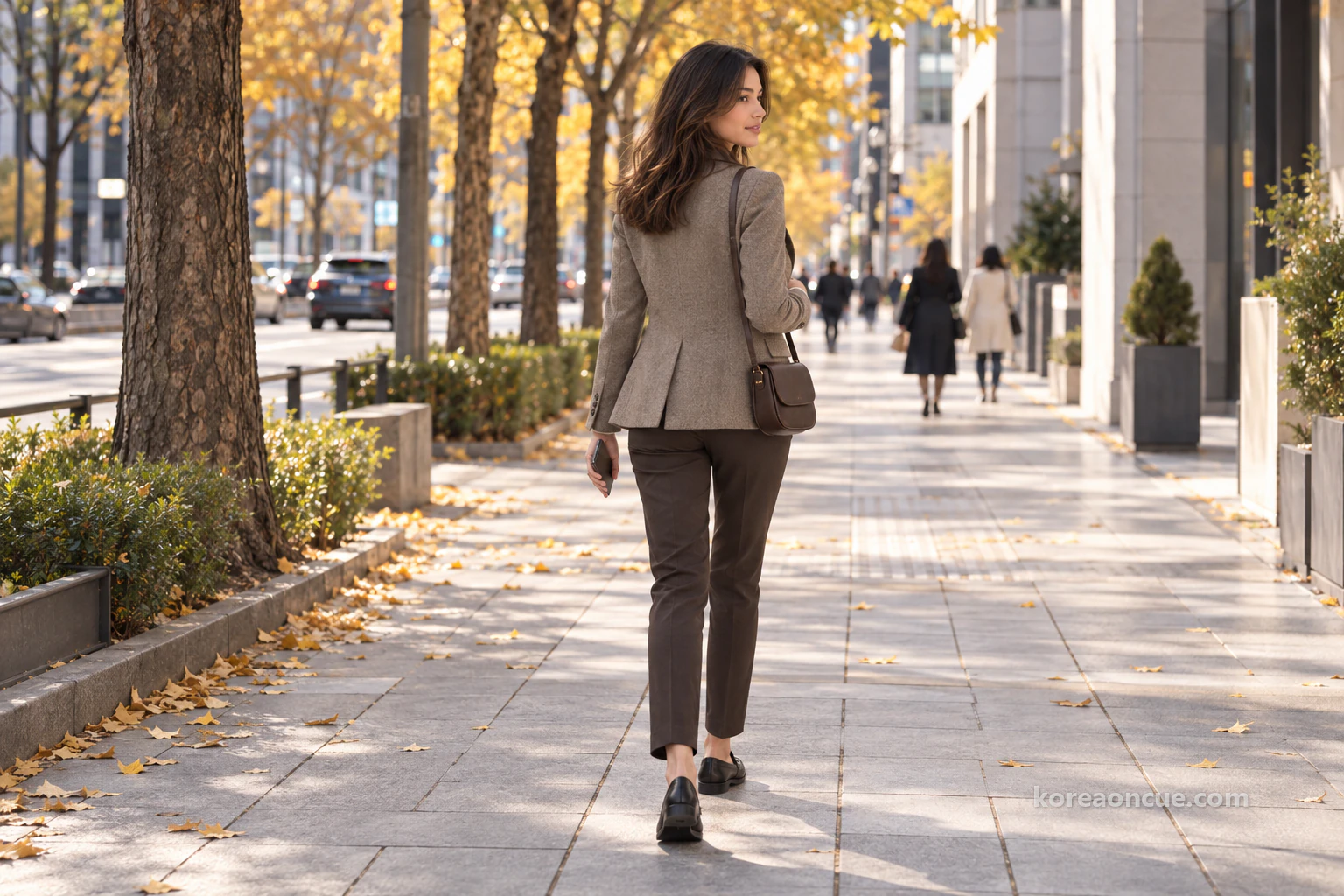 Elena seen at an angle from behind walking along a wide sidewalk lined with tall yellow street trees, her phone in one hand and a small bag on her shoulder, a broad road and buildings beside her. Editorial image.