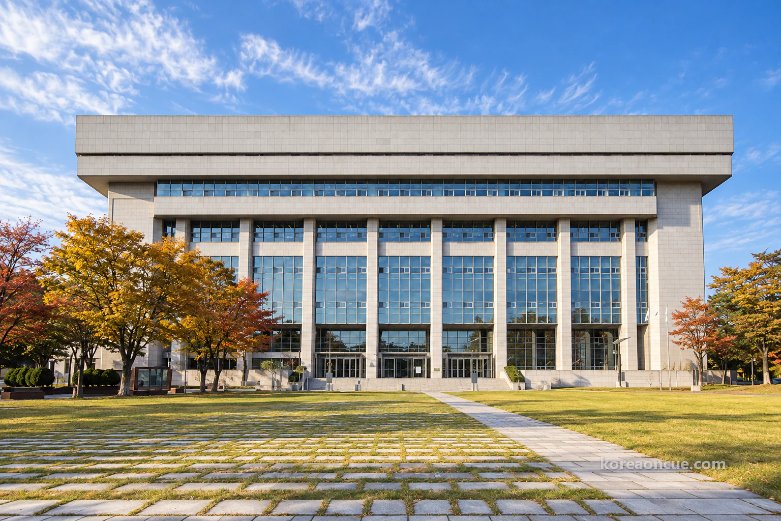 A wide, box-shaped building clad in pale grey stone seen straight on across a lawn on a clear late-autumn afternoon, a heavy grey band along the top, glass curtain walls between slim pillars from the ground floor to the fourth, square stone slabs set in the grass, a broad walkway and autumn trees on both sides. Editorial image.