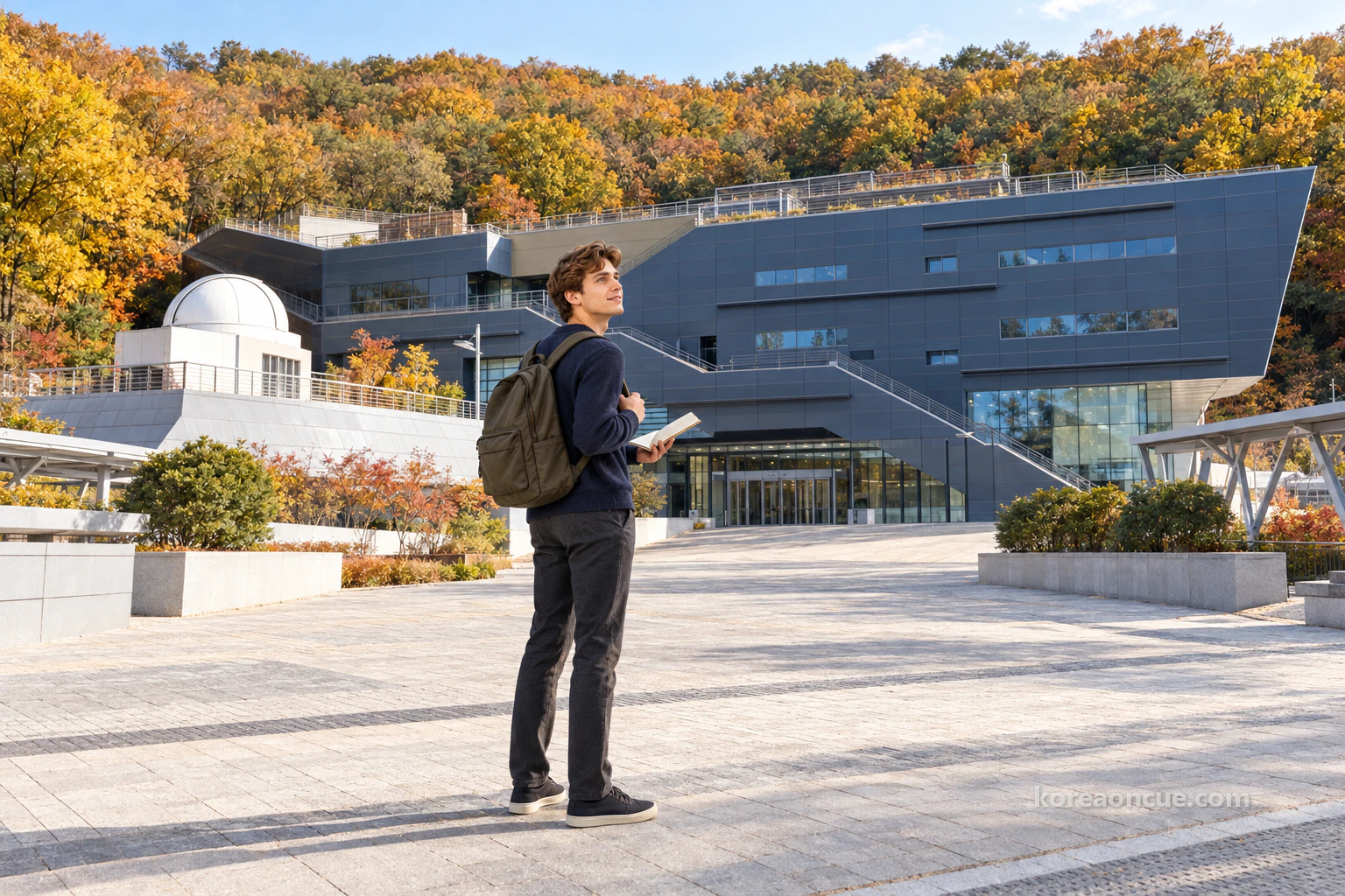 Noah seen at an angle from behind on a clear late-autumn morning, standing on a wide paved plaza and looking up at the upper floors of an angular three-storey building clad in dark blue-grey metal panels, with a hillside forest in yellow and orange behind it, an open notebook in one hand and his other thumb hooked under his backpack strap, a long glass entrance on the ground floor and a white observatory dome on an outdoor deck to the left of the building. Editorial image.