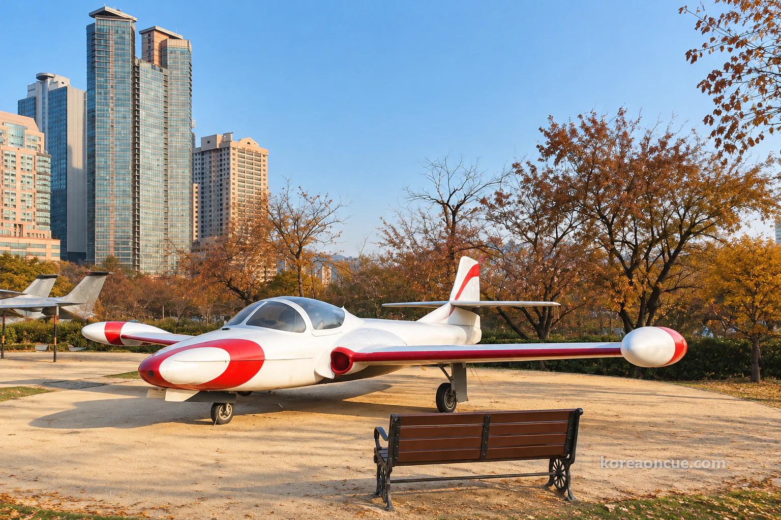 沒有人的晚秋晴朗早晨的 Air Park,沙色圓形廣場中央一架機鼻與翼尖漆成紅色的白色教練機,後方交疊的其他退役機體尾翼與機翼,廣場邊空著的木頭長椅,公園樹木後方的高樓公寓輪廓與寬闊天空。編輯示意圖。
