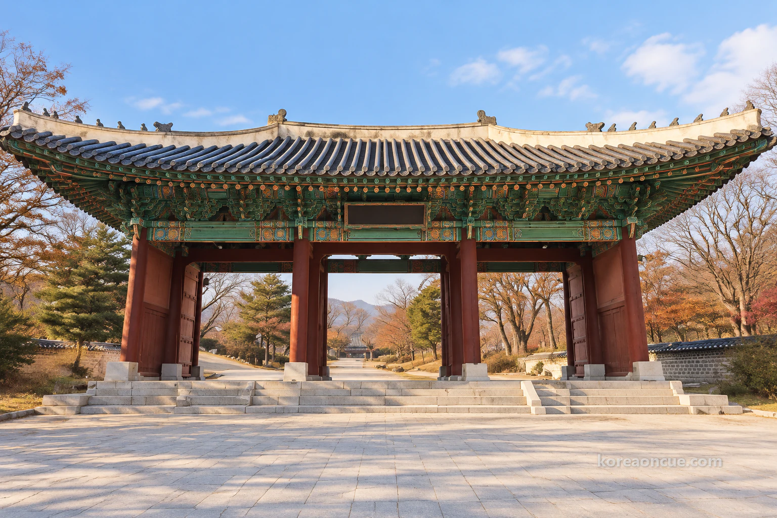 A palace main gate seen straight on across a broad stone-paved forecourt on a clear late-autumn afternoon, three open bays between four red pillars, blue-green dancheong under wide outspread eaves and a grey-tiled roof with gently upturned corners, a low stone base and short walls on either side, almost bare trees behind and the palace grounds softly visible through the bays. Editorial image.