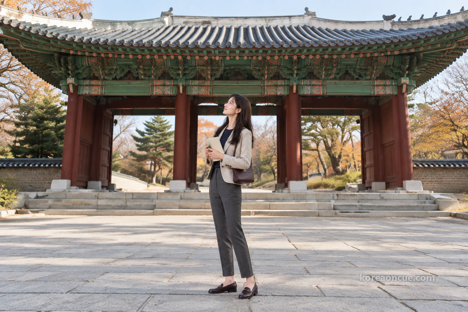 Rina seen at an angle from behind on a clear late-autumn afternoon, standing between two red pillars of the main gate and tilting her head back to look up under the eaves, both arms holding a closed notebook to her chest, blue-green dancheong and wide eaves above her, stone paving under her feet and short walls on either side. Editorial image.