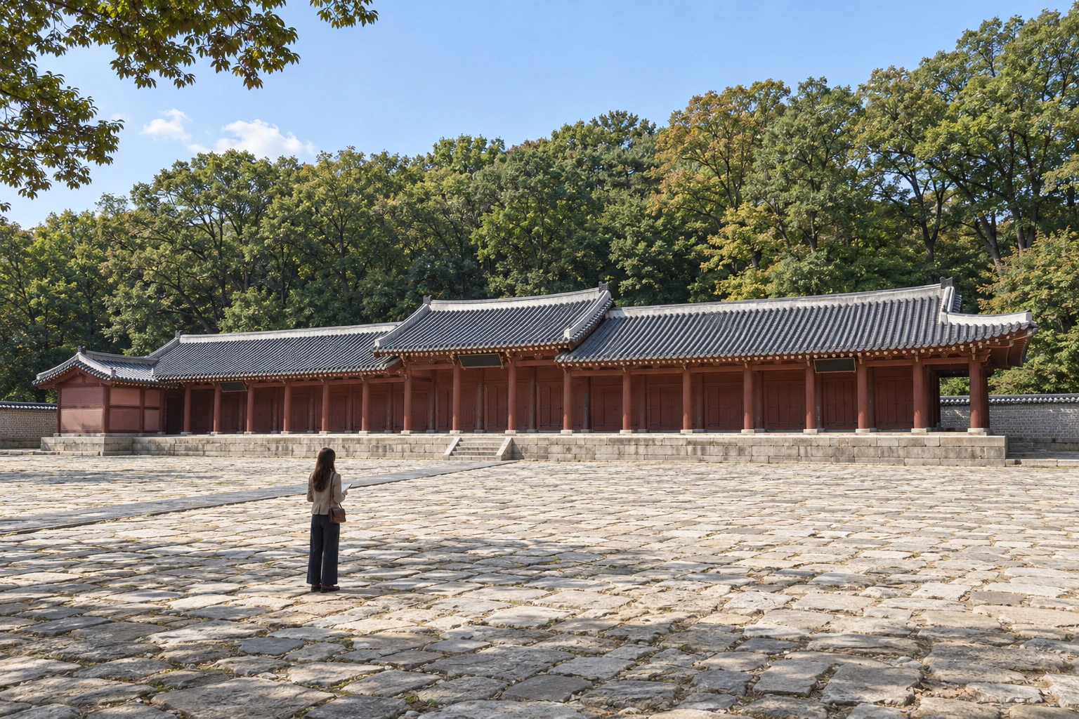 숲에 둘러싸인 긴 단층 건물 앞 넓은 돌 마당에 아주 작게 선 여성의 뒷모습 원경. 편집 이미지.