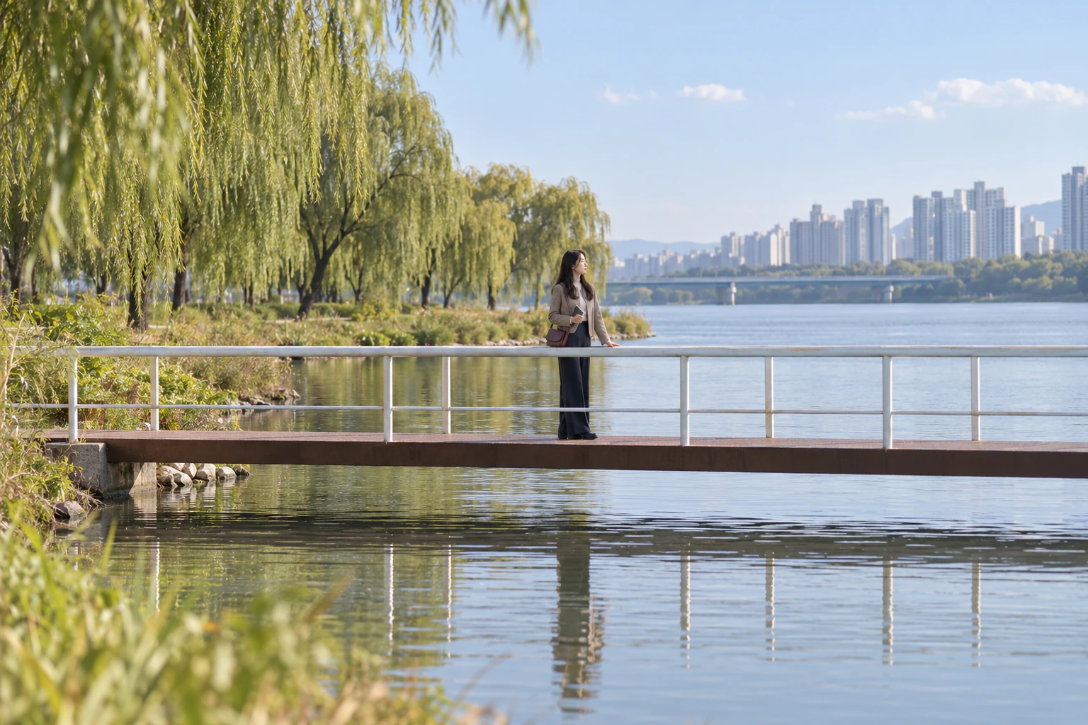 A short footbridge seen side-on from low down by the water. Halfway across, a woman stands with a hand on the railing, looking along the water, her full-length side view reflected in the calm surface. Face not shown in detail. Editorial image.