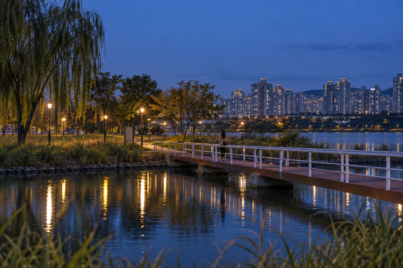 Blue evening, a wide view from the islet towards the park. On a low bridge across calm water, a very small woman walks away towards the park; the park’s path lamps and distant windows are lit. Editorial image.