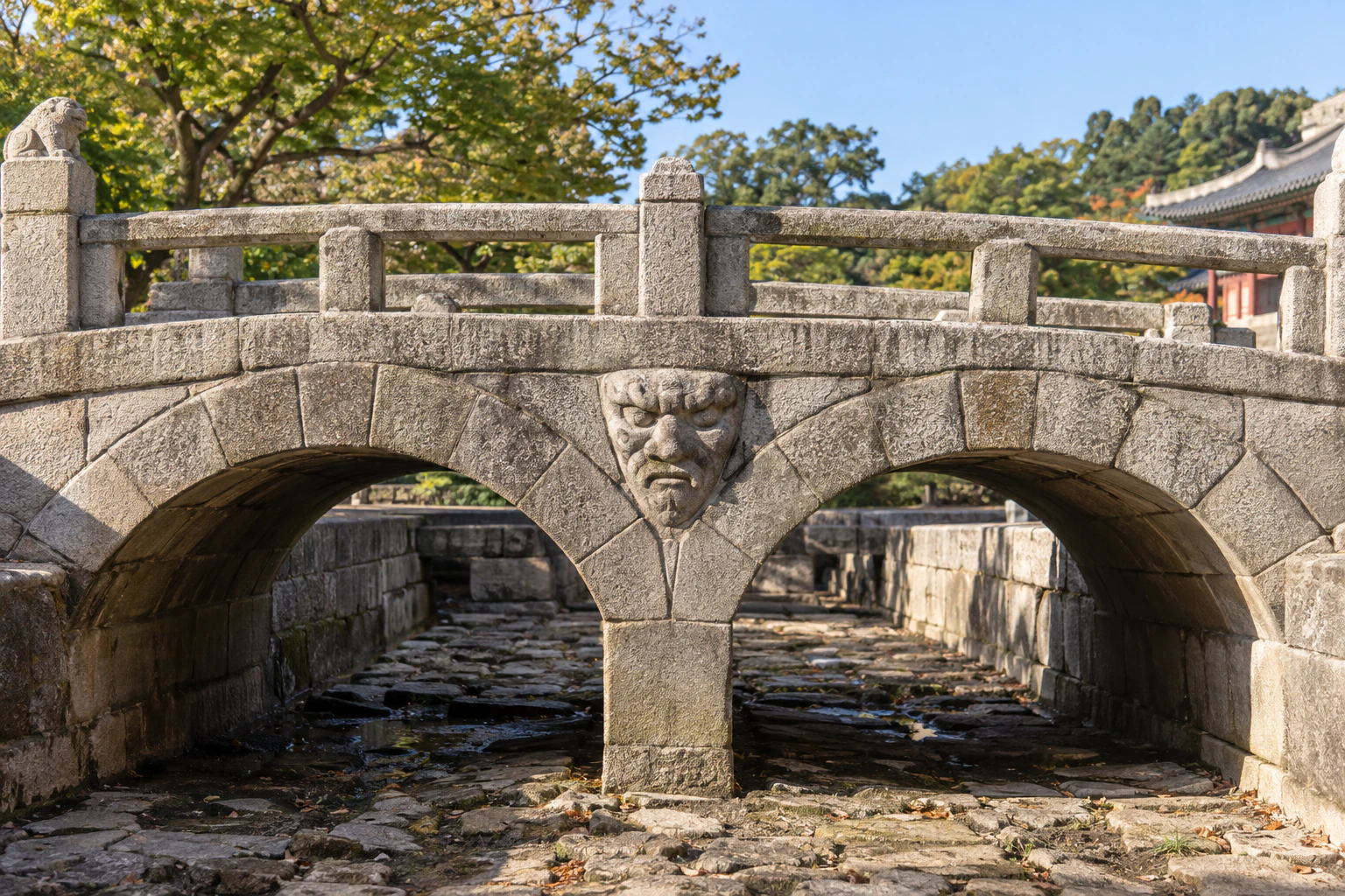 Two semicircular arches under a stone bridge with a broad-browed goblin face carved between them, above a stone-lined channel. No people. Editorial image.
