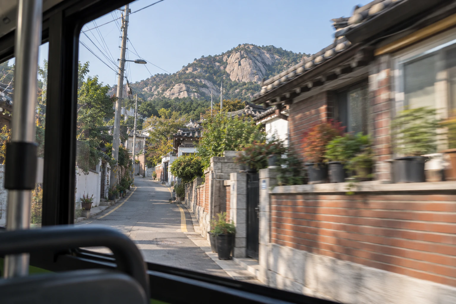 버스 창틀 너머로 낮은 집과 담장이 지나가고, 지붕 위로 바위가 드러난 산이 보인다. 문자는 넣지 않음. 편집 이미지.